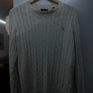 Beige stickad tröja från GANT - Säljer en snygg beige stickad tröja från GANT i premium bomull. Den har ett klassiskt flätmönster och rund halsringning. Perfekt för både höst och vinter, och den är i mycket bra skick. Passar till både vardag och lite finare tillfällen.