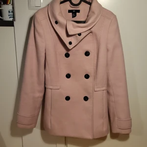 Rosa kappa från H&M - Säljer en superfin rosa kappa från H&M i storlek 40. Den har en dubbelknäppt design med svarta knappar och en stilren krage. Perfekt för höst och vår, och ger en elegant touch till vilken outfit som helst. Kappan är i mycket bra skick och redo för nya äventyr!