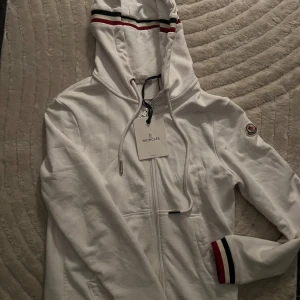 Vit hoodie från Moncler S M - Säljer en snygg vit hoodie från Moncler i mycket bra skick. Den har en dragkedja framtill och klassiska Moncler-ränder i rött och blått på huvan och ärmsluten. Små prickar fram på tröjan som bilden visar, därav priset! Använd en gång så i jätte bra skick annars! Obehandlad prickar. Jätte fin tröja. Passar S och M