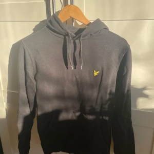 Svart Lyle & Scott hoodie i bra skick - Säljer en snygg svart hoodie från Lyle & Scott i mycket bra skick. Den har en gul broderad logga på bröstet och en praktisk magficka. Perfekt för höst och vinter, och passar både till vardags och chillkvällar. Storleken är normal och materialet är mjukt och bekvämt.