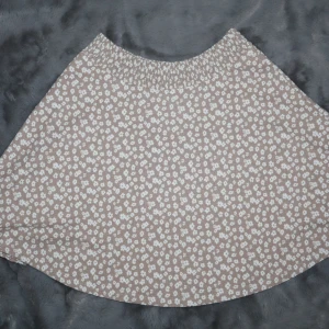 Beige blommig kjol - Beige kjol med ett blommigt mönster i vitt. Den har en elastisk midja. Kjolen är använd vid enstaka tillfälle!