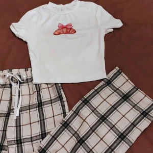 Pyjama set 🎀 🥐 - Säljer en superfin vit croppad t-shirt med ett gulligt croissant-tryck med rosa rosett på bröstet. 🎀 🥐 Perfekt för en avslappnad dag eller en mysig fika. T-shirten är kortärmad. Passar perfekt till  dessa rutiga byxor! ✨ (Båda delarna är nya)