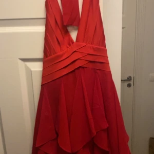 Röd klänning med volanger - Snygg röd klänning med volanger och halterneck-design. Perfekt för festliga tillfällen! Den har en dragkedja i sidan för enkel påklädning och är i mycket bra skick. Klänningen är i en härlig röd färg som verkligen sticker ut.