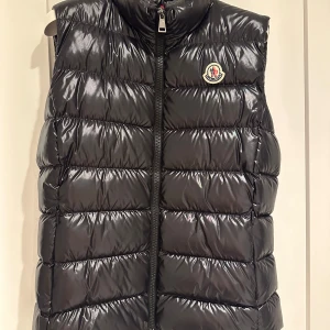 Svart dunväst från Moncler - Säljer en snygg svart äkta väst från Moncler i mycket bra skick. Västen har en dragkedja framtill och Monclers logga på bröstet. Perfekt för höst och vinter, håller dig varm och stilren. Passar både till vardags och lite finare tillfällen.finns kvitto och prislapp.