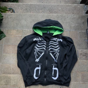 Skeletthoodie från NO FEAR - Cool svart hoodie från no fear med ett unikt skelettmönster på framsidan och en grön insida i huvan. Perfekt för höst och vår, med dragkedja och långa ärmar. Storleken är XXL men passar som M/L. Perfekt för dig som vill sticka ut lite extra!