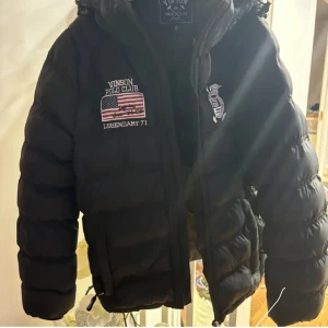 Svart pufferjacka från Vinson Polo Club - Säljer en svart pufferjacka från Vinson Polo Club i mycket bra skick. Jackan har broderade detaljer med amerikansk flagga och texten 'Legendary 71'. Den är perfekt för vintern med sin varma och vadderade design. Dragkedja framtill och justerbara snören i nederkant för bästa passform.