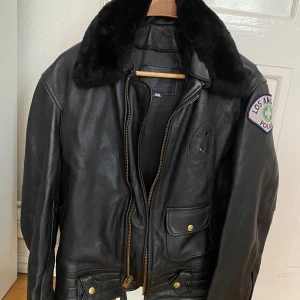 Original LAPD fodrat skinnjacka - Säljer en cool fodrat svart skinnjacka med pälsdetaljer på kragen.Jackan har en Los Angeles Police-patch på ärmen och stängs med dragkedja och knappar. Perfekt för höst och vinter, och ger en tuff look. Passar bra till både vardag och fest!