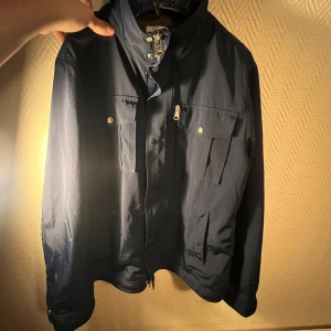 Mörkblå j.lindeberg field jacket - Säljer en snygg mörkblå jacka i bra skick. Den har en dragkedja framtill och knappar på fickorna. Perfekt för höst och vår, med en stilren look som passar till det mesta. Jackan har långa ärmar och en normal passform. Perfekt för dig som vill ha en stilren och praktisk jacka, som även är väldigt stilren och passar till mycket. Väldigt fint skick och skickar gärna fler bilder om det önskas