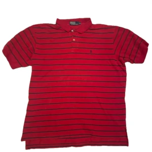 Röd randig pikétröja från Ralph Lauren - Säljer en snygg röd pikétröja från Ralph Lauren med svarta ränder. Tröjan har en klassisk krage och knappar framtill. Perfekt för både vardag och festliga tillfällen. Den är i mycket bra skick och redo för en ny ägare!