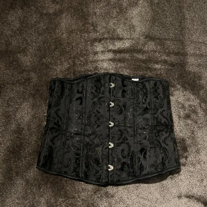 Svart korsett med brokadmönster - Säljer en elegant svart korsett med vackert brokadmönster. Den har hakknäppning fram och snörning bak för perfekt passform. Perfekt för festliga tillfällen eller som en stilfull accessoar. Använd endast en gång, så alltså i mycket gott skick❣️Storlek M men sitter perfekt på mig som vanligtvis har S