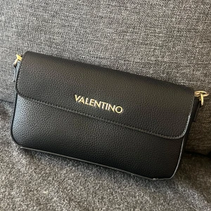 Svart axelväska från Valentino - Snygg svart axelväska från Valentino i skinn. Väskan har en elegant design med guldfärgade detaljer och en justerbar axelrem med Valentino-logga. Perfekt för både vardag och fest!