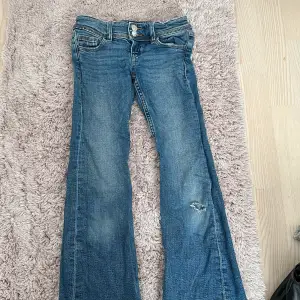 Säljer ett par blå bootcut jeans i bra skick. De har en snygg slitning på ena benet och dubbla knappar i midjan. Perfekta för en avslappnad stil med en vintage touch. Passar bra till både vardag och fest!. Priset kan diskuteras 