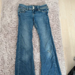 Blå bootcut jeans - Säljer ett par blå bootcut jeans i bra skick. De har en snygg slitning på ena benet och dubbla knappar i midjan. Perfekta för en avslappnad stil med en vintage touch. Passar bra till både vardag och fest!. Priset kan diskuteras 