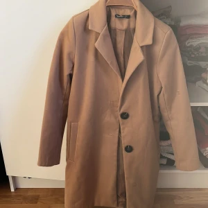 Beige kappa från Boohoo - Säljer en stilren beige kappa från Boohoo i storlek 34. Den har en klassisk design med stora knappar och långa ärmar. Perfekt för höst och vår, och ger en elegant touch till vilken outfit som helst. Kappan är i mycket bra skick och använd ett fåtal gånger