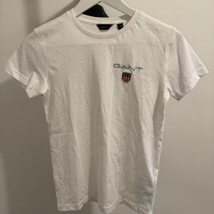 Vit t-shirt från GANT - Säljer en klassisk vit t-shirt från GANT i mycket bra skick. Den har en broderad logga på bröstet med ett stilrent emblem. Perfekt för en avslappnad och stilren look. Passar till alla tillfällen!
