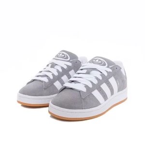 Grå och vita sneakers från Adidas - Snygga grå sneakers från Adidas med vita detaljer och klassiska ränder. De har en bekväm passform och är perfekta för vardagsbruk. Skorna har en gummisula i en ljusbrun nyans som ger bra grepp. Perfekta för både vår och höst!