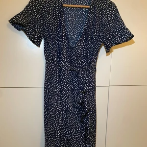 MARINBLÅ PRICKIG KLÄNNING  - Säljer en superfin mörkblå klänning med vita prickar. Den här v-ringad, är omlott (knyter i midjan) & har volang detaljer längst nere och slutet av ärmen. Fint skick. 