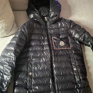 Säljer en snygg svart dunjacka från Moncler i mycket bra skick. Jackan har en glansig finish med dragkedja och tryckknappar. Den är långärmad och perfekt för kyliga dagar. Det finns en praktisk huva och en bröstficka med Moncler-logga. Perfekt för både höst och vinter! Om ni undrar något så kan ni skriva. Det går att se att den är äkta. Pris kan diskuteras 