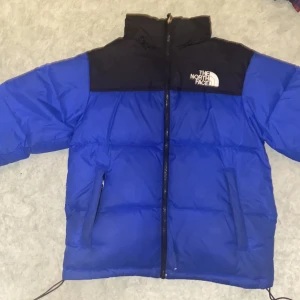 Blå och svart dunjacka från The North Face - Säljer en snygg och varm blå och svart dunjacka från The North Face. Perfekt för kalla vinterdagar med sin pufferstil och långa ärmar. Jackan har en dragkedja framtill och justerbara snören i nederkant för bästa passform. Den är i mycket bra skick och redo för nya äventyr.