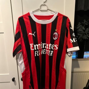 AC Milan fotbollströja från Puma - Säljer en AC Milan fotbollströja i rött och svart från Puma. Tröjan är kortärmad med klassiska ränder och har sponsorlogga på framsidan. Perfekt för fans som vill visa sitt stöd! 🖤❤️