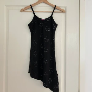 Svart asymmetrisk klänning med glitterdetaljer - Snygg svart klänning med asymmetrisk nederkant och glittriga blomdetaljer. Den är ärmlös med smala axelband och har en tight passform som framhäver figuren. Skulle passa någon bättre som är lite mindre än mig. 🎆🍸