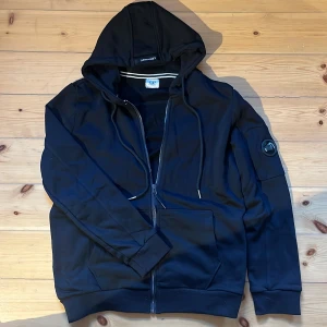 Svart hoodie zip från C.P. Company - Säljer en svart hoodie från C.P. Company som aldrig är använd⚫️ Den har en dragkedja framtill och en cool detalj med en logotyp på ärmen🔥Perfekt för höst och vinter, och den är långärmad med en slim/normal passform💪 Grymt skön och stilren! Den är i storlek M men är mycket mindre än så så jag skulle säga tt den är i storlek S🤩 Hör av dig vid frågor🙏