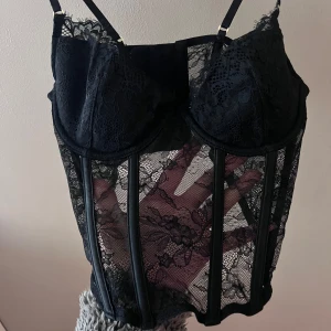 Svart spetskorsett - Snygg svart korsett i spets med justerbara axelband. Den har en genomskinlig design med vackra spetsdetaljer och ger en elegant look. Perfekt för en fest eller en speciell kväll. Den är i mycket bra skick och redo att bäras!