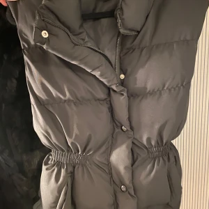 Svart dunväst från Dakfrogs - Säljer en svart dunväst från Dakfrogs i storlek M. Västen har en dragkedja och knappar framtill samt en elastisk midja för bättre passform. Perfekt för kyliga dagar och ger en stilren look.