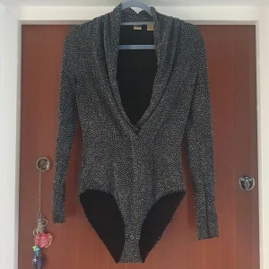 Svart och vit prickig bodysuit - Snygg svart bodysuit med glittriga prickar och djup v-ringning. Den har långa ärmar och en åtsittande passform, perfekt för en utekväll eller fest. Materialet är stretchigt, vilket gör den bekväm att bära hela dagen eller natten.