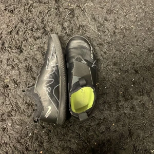Svarta Nike fotbollsskor - Säljer ett par svarta Nike fotbollsskor i bra skick. De har en cool design med vita detaljer och en slitstark sula. Perfekta för både träning och match! Skorna har en elastisk passform utan snörning, vilket gör dem lätta att ta på och av. Innersulan är grön och ger bra komfort. Skorna är perfelta nu inför vintern och futsal säsongen. Skorna kostar nypris 649kr.