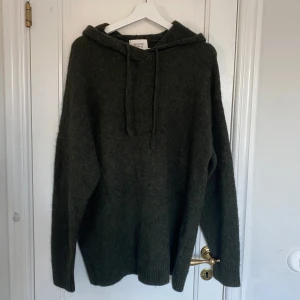 Mörkgrön stickad hoodie  - Säljer en supermysig mörkgrön hoodie från Second Female. Den är i ullmaterial och perfekt för kyliga dagar. Tröjan har en avslappnad passform med långa ärmar och en klassisk huva med dragsko. Perfekt för höst och vinter!