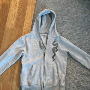 Säljer en snygg grå hoodie från Trapper i storlek M. Den har ett coolt ormmönster i svart och glittrande detaljer på framsidan. Perfekt för en avslappnad stil med dragkedja och huva. Passar bra till både vardag och chill.