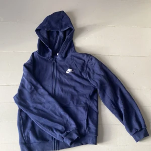 Mörkblå hoodie från Nike - Säljer en snygg och bekväm mörkblå hoodie från Nike i storlek 158-170cl. Den har en dragkedja framtill och en klassisk Nike-logga på bröstet. Oanvänd.