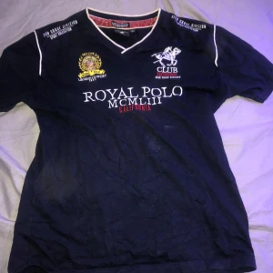 Svart t-shirt från Geographical Norway - Säljer en svart t-shirt från Geographical Norway med vit text och broderade detaljer. Den har en v-ringad hals och korta ärmar. På framsidan står det 'Royal Polo' och på baksidan finns en stor siffra 5. Perfekt för en casual look!