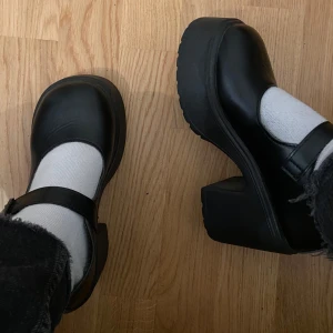Svarta Mary Jane-plattformsskor - Säljer ett par svarta Mary Jane-plattformsskor i syntetmaterial. De har en chunky klack och en justerbar rem över foten. Perfekta för att ge lite extra höjd och stil till din outfit. Skorna är i mycket bra skick och passar både till vardags och fest. Endast använda en kväll!