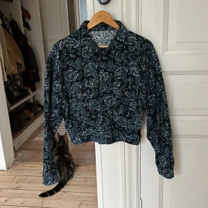 Blommig jacka i bomull - Säljer en superfin blommig jacka i 100% bomull, perfekt för vår och sommar. Den har ett snyggt blommönster i grått och blått och är i mycket bra skick. Jackan har långa ärmar och knappar framtill. Perfekt för att ge din outfit en unik touch!