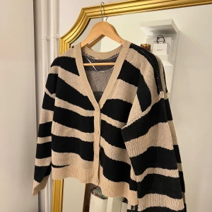 Randig kofta i svart och beige - Säljer en superfin randig kofta i svart och beige. Den har en avslappnad passform med långa ärmar och en djup v-ringning. Perfekt för höst och vår när du vill ha något mysigt men ändå stilrent. Koftan är i bra skick och passar perfekt till både jeans och kjol.