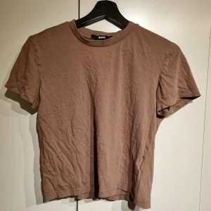 Brun t-shirt från bikbok - Säljer en enkel och stilren brun t-shirt från Bikbok. Den är kortärmad och har en normal passform, perfekt för vardagsbruk. Materialet känns mjukt och bekvämt, vilket gör den idealisk för både sommar och vår. Passar bra till jeans eller shorts för en avslappnad look.