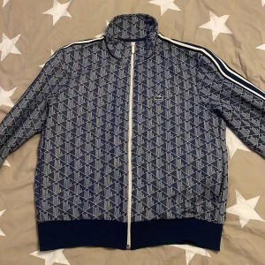 Lacoste  - Säljer en snygg mönstrad jacka från Lacoste i blått och vitt. Jackan har en dragkedja framtill och klassisk Lacoste-logga på bröstet. Perfekt för en sportig look eller vardagsstil. Regular fit och bekväm att bära.