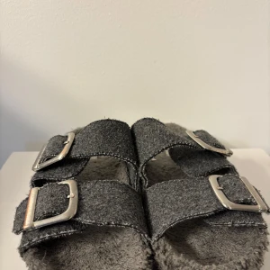 Grå inomhustofflor med spännen - Säljer ett par grå inomhustofflor i bra skick med dubbla spännen. De har en mjuk och fluffig insida som gör dem superbekväma att ha på sig. Perfekta för en avslappnad dag hemma eller en promenad i parken. Sulan är i bra skick med bra grepp. Billiga!