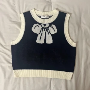 Mörkblå stickad väst med rosett - Säljer en superfin mörkblå stickad väst från Shein med en stor vit rosett på framsidan. Västen har vita ribbade kanter vid ärmhål och nederkant. Perfekt för lager-på-lager-stil under höst och vår!