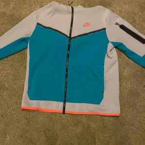 Grå och blå jacka från Nike - Säljer en snygg grå och blå hoodie från Nike. Den har en dragkedja framtill och en cool ficka med dragkedja på ärmen. Perfekt för höst och vår! Jackan har en orange kant nedtill som ger en extra pop av färg. Passar perfekt för träning eller vardagsbruk.