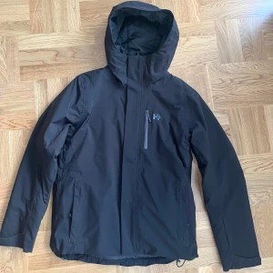 Svart Men's Panorama Jacket från Helly Hansen - Säljer en svart Helly Hansen Men's Panorama Jacket (ORGINAL PRIS 3300kr) i mycket bra skick som jag köpte för Skid säsong men inte använde särskilt mycket då jag hade två stycken jackor. Jackan är perfekt för vinter och skidåkning med sitt slitstarka material och stilrena design. Den har en dragkedja kardboard, snöskydd, flertal fickor, bland annat fick för liftkort. Skriv gärna för fler bilder!