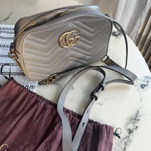 Grå quiltad cross body väska  - Köpt här på Plick men onvänd. Ren men har små repor (på guld bokstäverna och undersidan) som syns på bilderna. Fin grå färg. 