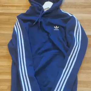 Säljer en snygg blå hoodie från Adidas med de klassiska tre ränderna längs ärmarna. Perfekt för både träning och vardag. Den har en känguruficka och justerbar huva med dragsko. Adidas-loggan är broderad på bröstet. Superbekväm och stilren!