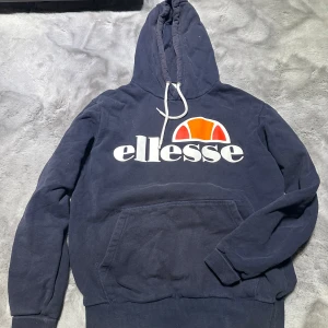 Mörkblå hoodie från Ellesse - Säljer en snygg mörkblå hoodie från Ellesse med deras ikoniska logga i vitt och orange på bröstet. Den har en stor ficka fram och justerbar huva med snören. Perfekt för en avslappnad stil eller träning. Hör av dig vid intresse!
