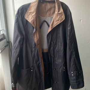 Blå vindjacka från Atelier Création - Snygg blå vind jacka från Atelier Création med brunt foder och krage. Jackan  har knappar och dragkedja, samt praktiska fickor med knappar. Perfekt för höst och vår!