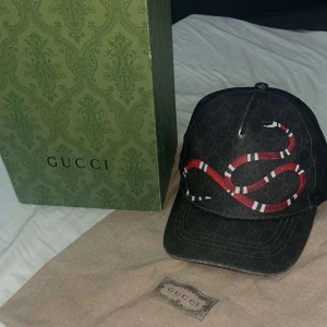 Svart keps med orm från Gucci - Säljer en snygg svart keps från Gucci med en cool orm design i rött och vitt. Kepsen är i mycket bra skick och har en justerbar passform. Perfekt för att ge din outfit en lyxig touch!