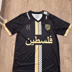 Palestina fotbollströja 24/25 - Säljer en snygg svart fotbollströja med guldiga detaljer och ett unikt mönster. Tröjan har korta ärmar och ett coolt tryck på framsidan. Perfekt för dig som vill sticka ut på planen eller bara ha en stilren look. Helt ny med taggar kvar! Finns i storlek S och M!