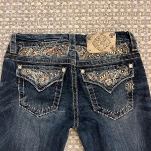 Blå jeans med broderade detaljer från Miss Me - Snygga blå jeans från Miss Me med unika broderade detaljer och glittriga stenar på bakfickorna. De har en bootcut-stil och är perfekt för att ge en extra touch till din outfit. Jeansen är i bra skick och har en lågmidjad passform.🩷 Jag är 165cm lång och de är storlek 27 i midjan🩷